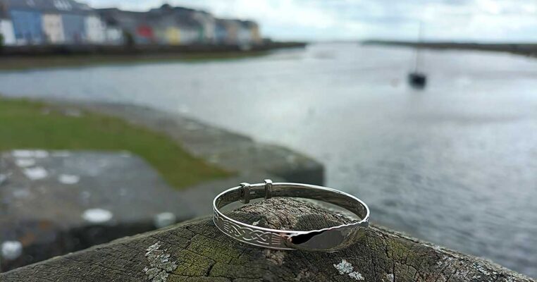 Engravable hallmarked sterling silver Claddagh baby bangle for Christening and new baby gift, with free personalised engraving and tracked delivery. Photographed at the Claddagh, Galway, Ireland with the River Corrib, the Long Walk and a traditional Galway Hooker sailboat in the background.