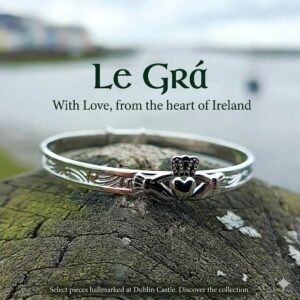 Sterling silver adjustable Claddagh baby bangle with traditional hands, heart, and crown design, featuring Celtic scroll engraving, set against a blurred Irish coastal background.