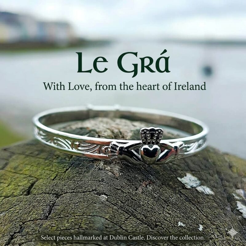 Sterling silver adjustable Claddagh baby bangle with traditional hands, heart, and crown design, featuring Celtic scroll engraving, set against a blurred Irish coastal background.