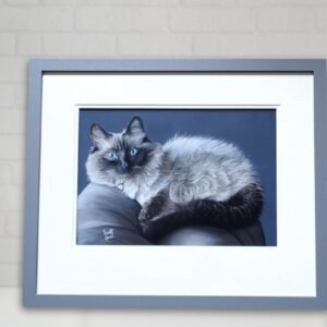 Framed painting of a fluffy cat with blue eyes, lounging on a gray surface, displayed on a white shelf next to a white vase with orange flowers.