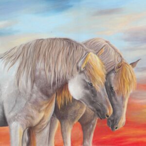 Two horses with light brown manes stand close together, heads bowed gently, against a vibrant sky blending blue, orange, and red hues, suggesting sunrise or sunset.