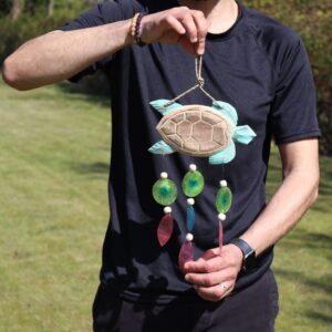 Handcrafted aqua turtle driftwood windchime with capiz shell drops hanging in a garden setting. Soft coastal windchime design bringing relaxed summer ambience.