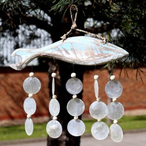 Bluewashed fish driftwood windchime with natural capiz shells hanging in a bright garden setting. A soft coastal touch for relaxed living spaces.